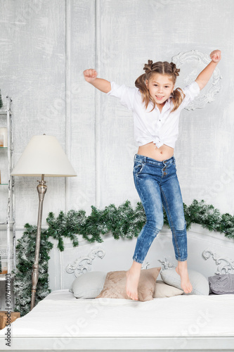 Fototapeta Naklejka Na Ścianę i Meble -  Little cute beautiful girl having fun and jumping on the bed in blue jeans and a white shirt on the background of a room in the New Year's decoration with a Christmas tree.