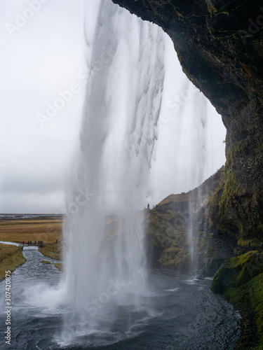 The impressive Seljalandsfoss waterfall seen from the wide cavern behind the water curtain. With the meadow and Seljalandsá river in the background. South Coast of Iceland.