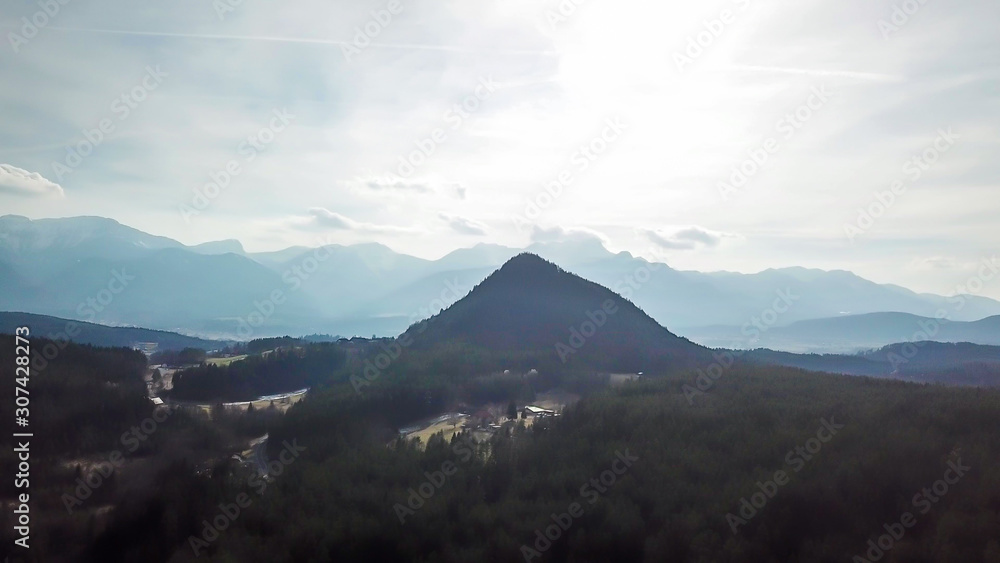 Areal, drone view on Kathreinkogel in Schiefling am See, Austria. The hill has a perfect pyramid shape. It is overgrown with forest. There are high Alps in the back. Little village under the hill.