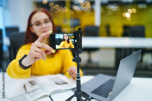 Hand woman holding camera for recording video while sitting at office. Teenager student in a yellow sweater with laptop having fun vlogging live feeds on social media. Technology and videoblog concept photo