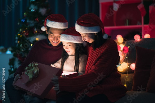 Young Asian family celebration in Christmas day, father mother and daughter siting on sofa and open gift box which exciting smiling and felling happy in living room at home at night time. Merry Xmas.