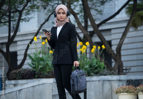 Asian Muslim woman walking while using headphone and carrying bag outdoor.