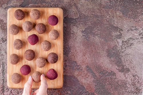 Woman Hand Take a Homemade Raw Vegan Cacao Energy Balls from Wooden Board on Marble Background. Healthy Sweets from Nuts and Dates. Concept of Natural Vegetarian Handmade Dessert. Top View. Copy Space