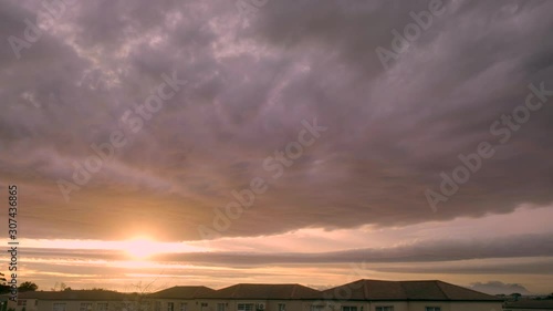 Sky filled with thick rain clouds moving over suburban roof tops as sun peeks through a break in the clouds in the distant with golden hour sunset in 4K. photo