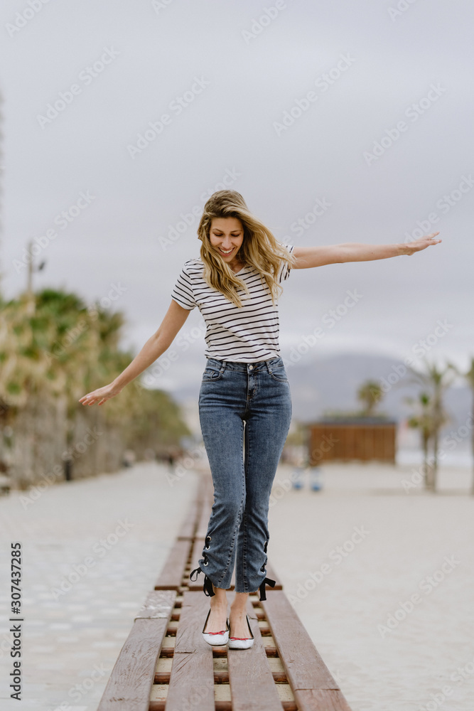 Happy cheerful female in casual clothes walking with arms spread on wooden bench and enjoying summer day at city seafront Happy cheerful female in casual clothes walking with arms spread on wooden bench and enjoying summer day at city seafront