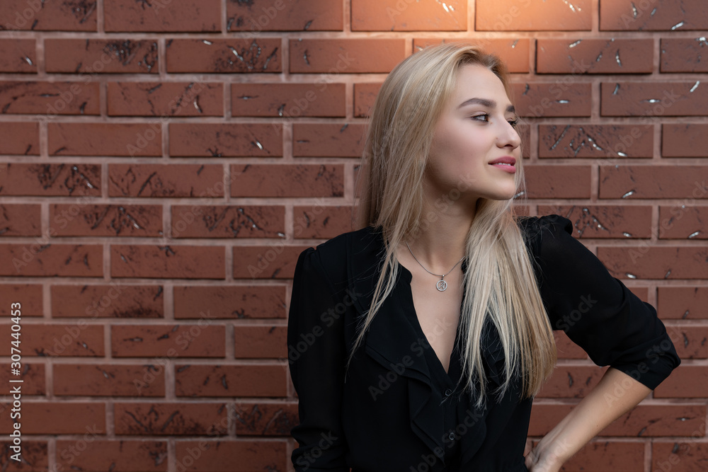 Attractive young woman with blonde hair and red lips looking at the right side, holding hand on her waist, red brick wall on the background