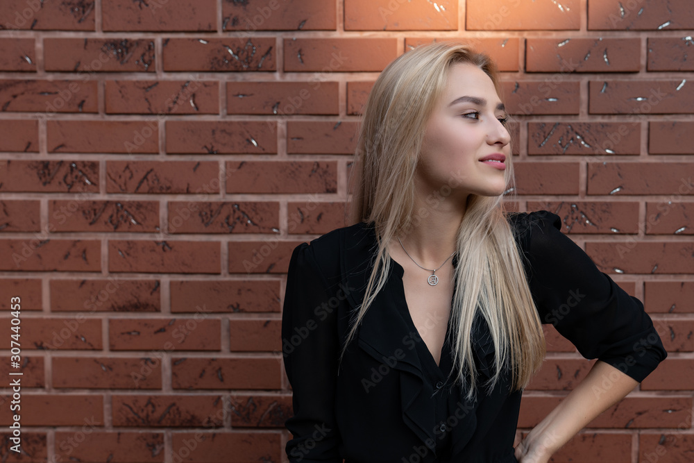 Attractive young woman with blonde hair and red lips looking at the right side, holding hand on her waist, red brick wall on the background Attractive young woman with blonde hair and red lips looking at the right side, holding hand on her waist, red brick wall on the background