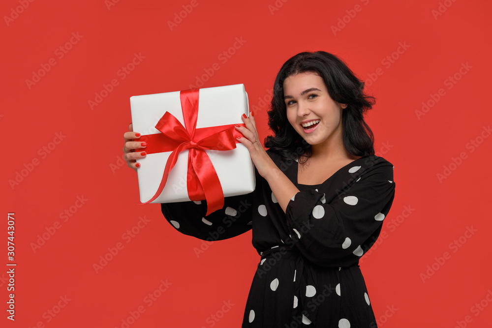 Brunette woman in a black dress smiling holding Christmas giftbox of white color with red ribbon. Brunette woman in a black dress smiling holding Christmas giftbox of white color with red ribbon.