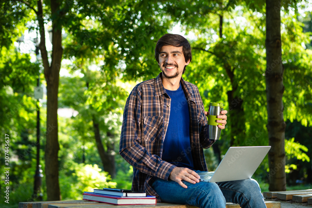 Fototapeta premium Young brunet freelancer with beard and mustache sitting with a laptop and coffee in a park.