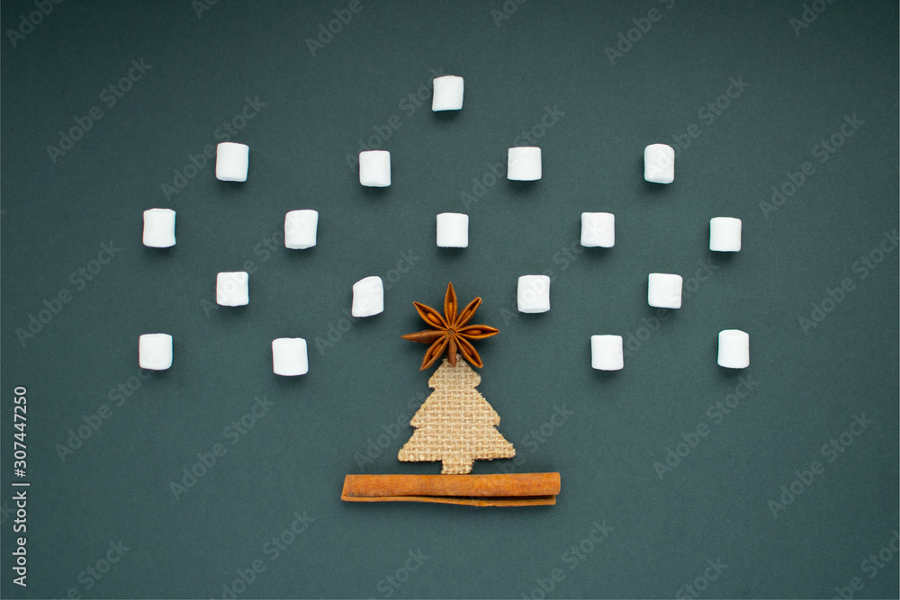 Creative minimalistic christmas flat lay. Marshmallow snowfall with one decorative fir tree, anise as star on christmas tree and cinnamon sticks as ground at green background. Nordic style.