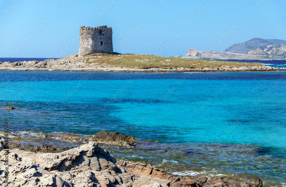 Aragonese (Torre della Pelosa) tower in Stintino, Sardinia, Italy. Landmark of Stintino on azure blue water and rocks, mountains in the background.