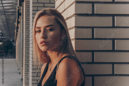 Sensual blonde girl with natural makeup dressed in black top leaned on the white brick wall and looks at the camera