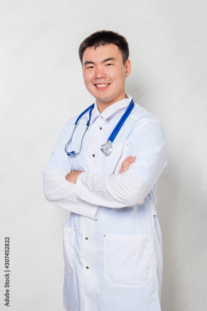 A young Asian doctor in a white coat, with a stethoscope on his neck stands holding his hands on his chest alone on a white background. A young Asian doctor in a white coat, with a stethoscope on his neck stands holding his hands on his chest alone on a white background.