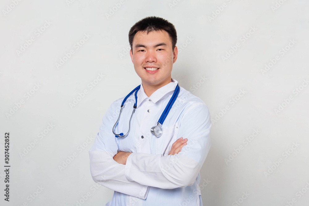 A young Asian doctor in a white coat, with a stethoscope on his neck stands holding his hands on his chest alone on a white background. A young Asian doctor in a white coat, with a stethoscope on his neck stands holding his hands on his chest alone on a white background.