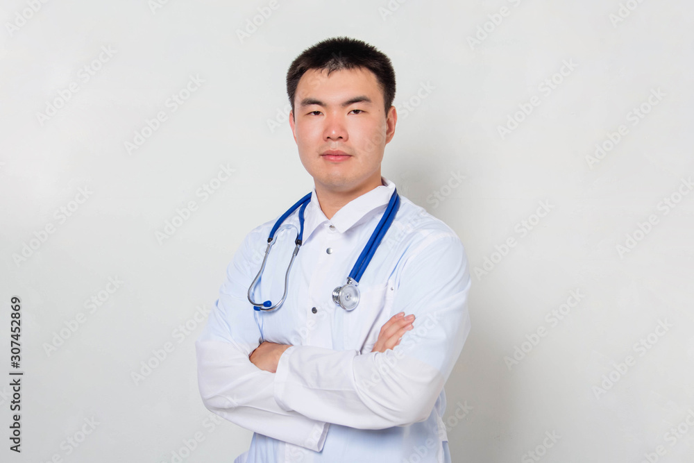 A young Asian doctor in a white coat, with a stethoscope on his neck stands holding his hands on his chest alone on a white background. A young Asian doctor in a white coat, with a stethoscope on his neck stands holding his hands on his chest alone on a white background.