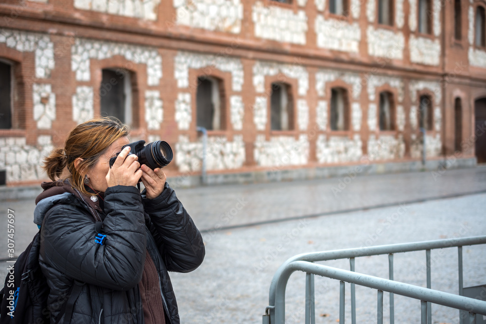 Mature woman making a photo, dressed casual, winter, with unfocused old building background, horizontal