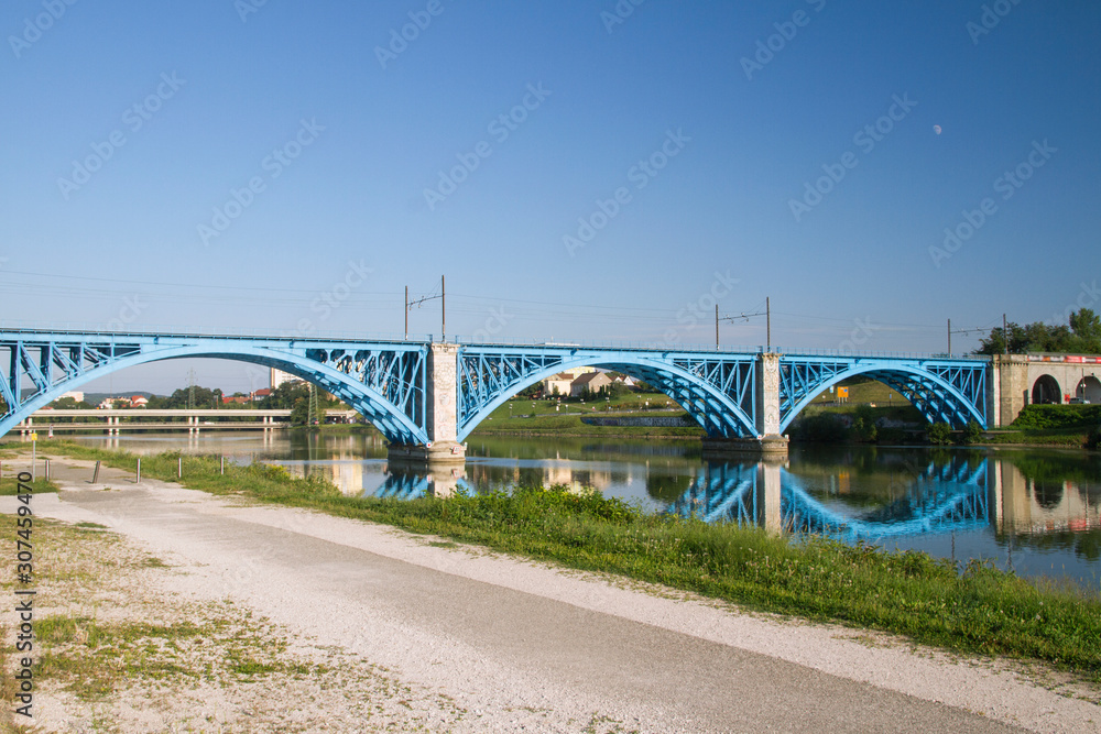 Promenade by Blue steel railway bridge over Drawa river. Reflections in water. Maribor, Slovenia.