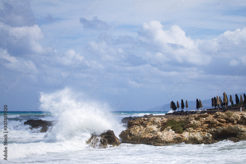 Splashing high wave on the rough sea. Folded umbrellas on empty closed beach because of bad weather. Cloudy sky on a windy day. Crete Island. Greece. Splashing high wave on the rough sea. Folded umbrellas on empty closed beach because of bad weather. Cloudy sky on a windy day. Crete Island. Greece.