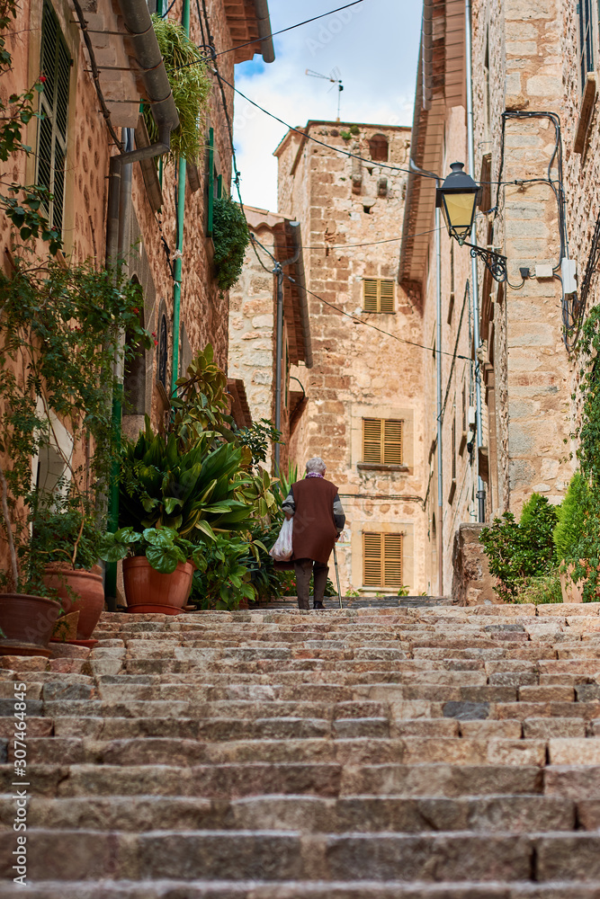 Elderly woman climbing stone stairs in Mallorcan village. Street with ornamental plants. Fornalutx, Mallorca