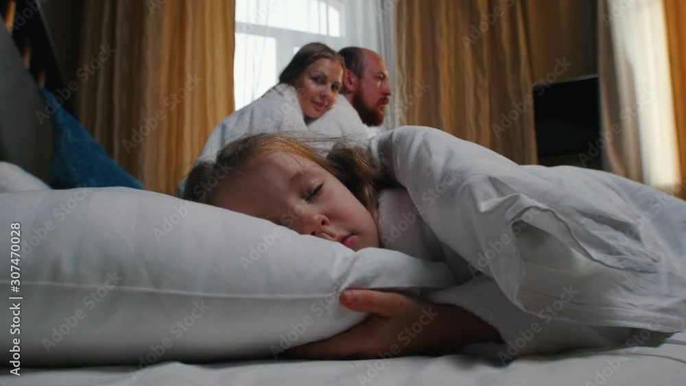 A family in the hotel room - a little girl sleeping in bed - her mom and dad looking at her A family in the hotel room - a little girl sleeping in bed - her mom and dad looking at her