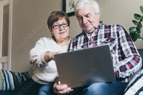 Elderly couple doing shoppings online on the sofa at home, looking at laptop screen and smiling