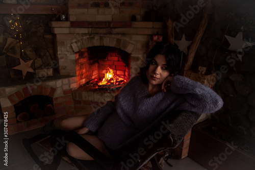 Portrait of pregnant woman wearing knitted warm sweater sitting in rocking chair against the burn fire in brick fireplace at home and holding a cup of tea