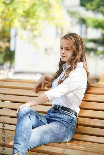  a girl in a white blouse and blue jeans sits on a wooden bench in autumn Park square, miss autumn © borisblik