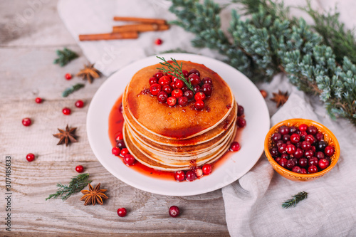 Stack of american pancakes with fresh cranberry and jam in white plate on wooden rustic table decorated Christmas tree, delicious dessert for breakfast in winter, vintage style. 