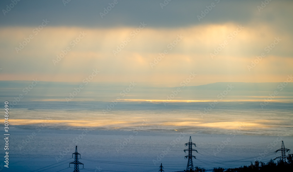 Cloudy sky, sunset is approaching, the rays of the sun fall on the winter sea. In the foreground are the towers of power lines. Cloudy sky, sunset is approaching, the rays of the sun fall on the winter sea. In the foreground are the towers of power lines.