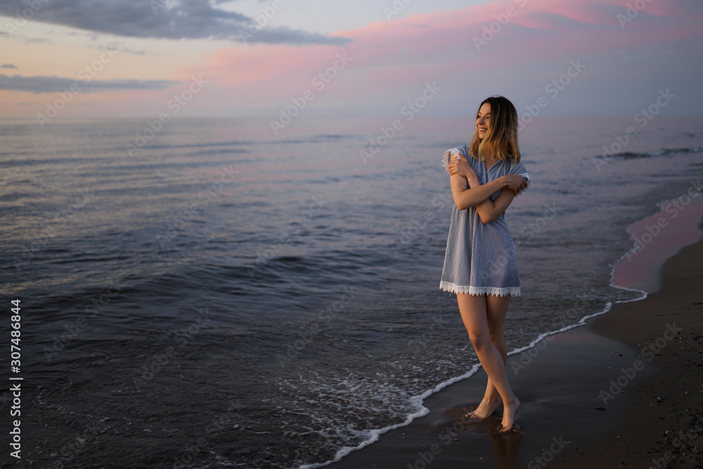 Standing full height: Portrait of a Beautiful blonde woman in a light blue dress on the Baltic Sea beach during sunset with vivid colors