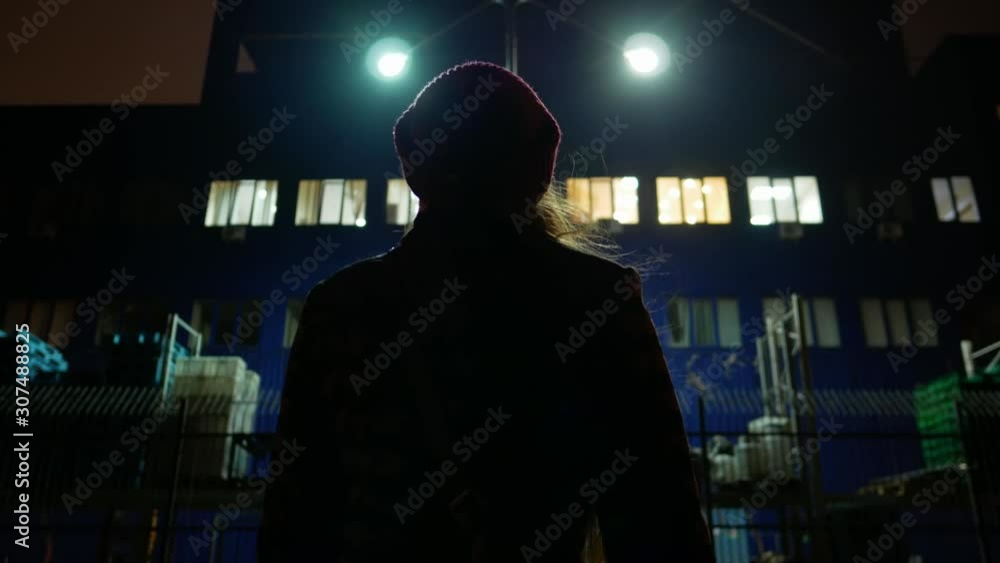 silhouette of a girl in a hat stands near a building fenced by a fence at night silhouette of a girl in a hat stands near a building fenced by a fence at night