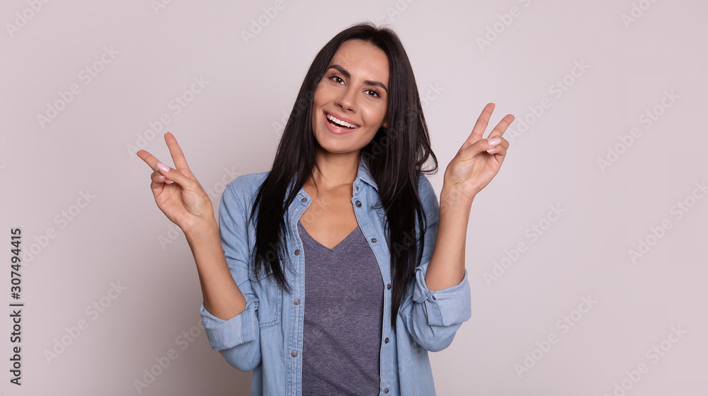 Love and peace. Close-up photo of a playful dark-haired girl in a casual outfit, who is looking in the camera with appealing smile and showing peace gesture with her hands. Love and peace. Close-up photo of a playful dark-haired girl in a casual outfit, who is looking in the camera with appealing smile and showing peace gesture with her hands.