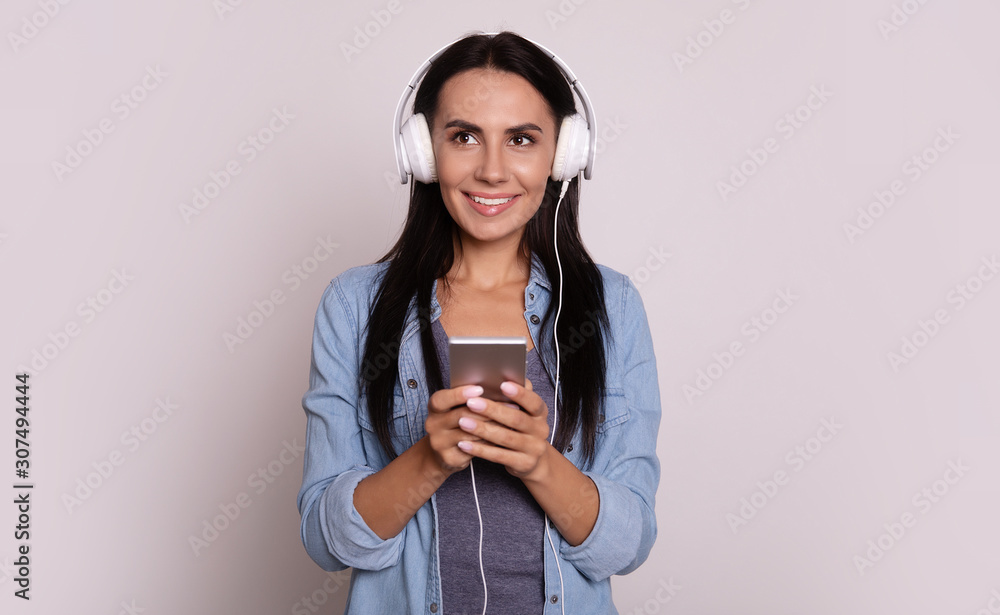 Cool beats. Close-up photo of wonderful young girl with long dark hair in big white headphones, who is looking to the left, smiling broadly and holding a smartphone. Cool beats. Close-up photo of wonderful young girl with long dark hair in big white headphones, who is looking to the left, smiling broadly and holding a smartphone.