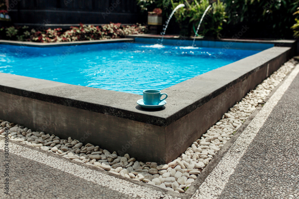 A blue cup of coffee stands on the corner of a tropical pool overlaid with white stone. Comfortable vacation in Asia