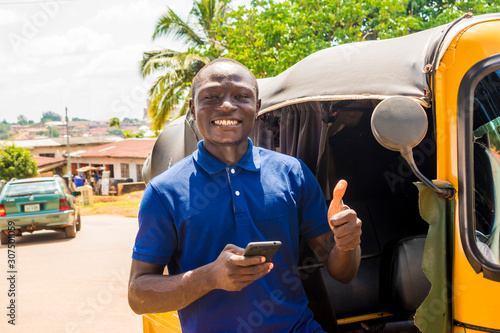 cheerful african man standing next to his tuk tuk taxi smiling and using his smart phone giving a thumbs up photo