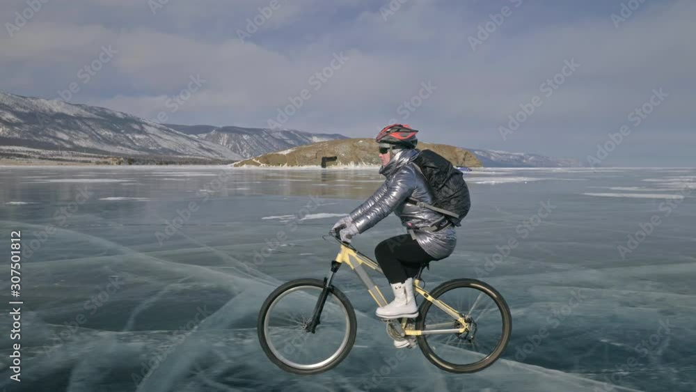 Woman is riding bicycle on the ice. The girl is dressed in a silvery down jacket, cycling backpack and helmet. Ice of the frozen Lake Baikal. The tires on the bicycle are covered with special spikes Woman is riding bicycle on the ice. The girl is dressed in a silvery down jacket, cycling backpack and helmet. Ice of the frozen Lake Baikal. The tires on the bicycle are covered with special spikes