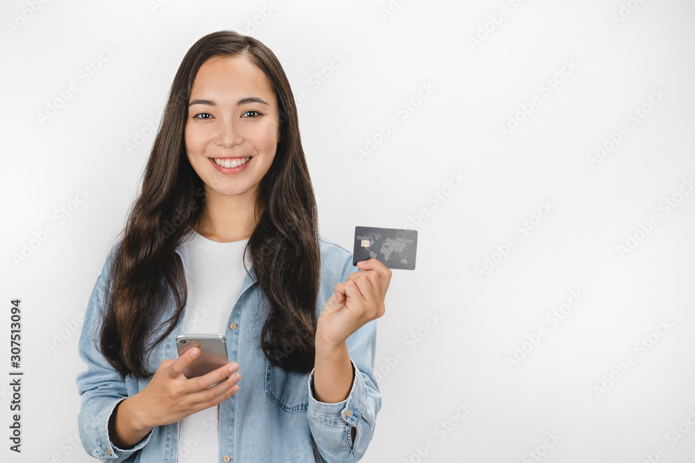 Portrait of smiling asian woman holding credit card and mobile phone while looking at camera isolated over white background Portrait of smiling asian woman holding credit card and mobile phone while looking at camera isolated over white background