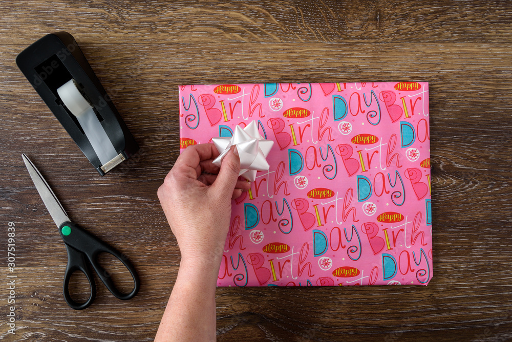 Gift wrapped with pink birthday paper, woman’s hand with white bow, tape dispenser and scissors, wood table Gift wrapped with pink birthday paper, woman’s hand with white bow, tape dispenser and scissors, wood table