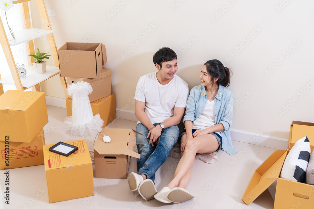 Young happy asian couple man and woman talking and sitting on the floor at new home. Couple sitting with carton package box storage to move in empty home. Young married asian relocating home concept. Young happy asian couple man and woman talking and sitting on the floor at new home. Couple sitting with carton package box storage to move in empty home. Young married asian relocating home concept.