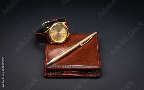 Brown leather wallet with watch and gold pen isolated on black gradient background. Luxury accessories. Gift concept for men. 