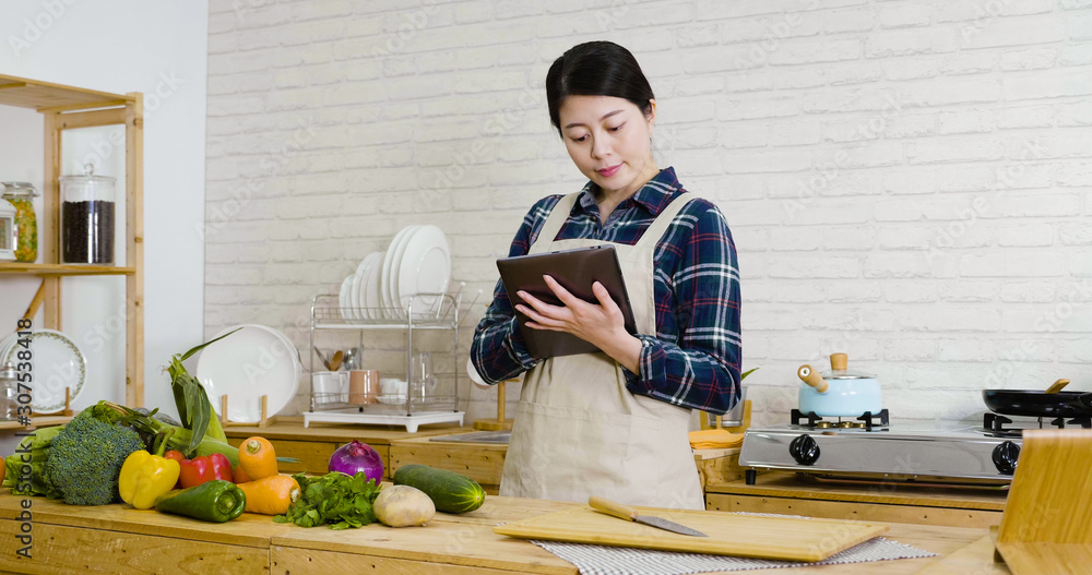 young asian japanese girl in apron using digital tablet device while cooking in kitchen at home. happy smiling woman preparing healthy meal and searching vegan recipes via portable computer indoors. young asian japanese girl in apron using digital tablet device while cooking in kitchen at home. happy smiling woman preparing healthy meal and searching vegan recipes via portable computer indoors.