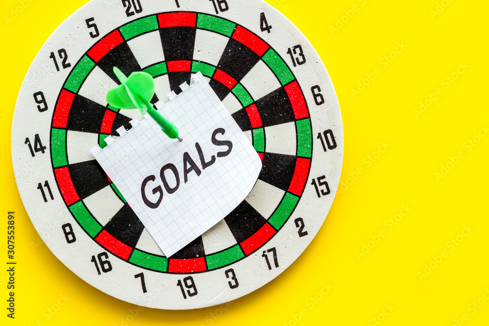 Goals text attached by dart to dart board on yellow background top-down copy space Goals text attached by dart to dart board on yellow background top-down copy space