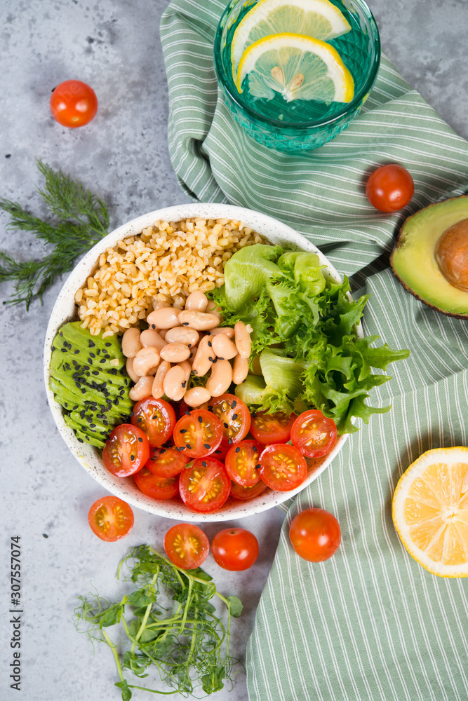 A plate of fresh salad with white beans, bulgur, cherry tomatoes and avocado, decorated with black sesame seeds with products around the plate. Vertical photo, top view A plate of fresh salad with white beans, bulgur, cherry tomatoes and avocado, decorated with black sesame seeds with products around the plate. Vertical photo, top view
