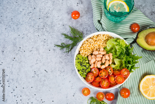 A plate of fresh salad with white beans, bulgur, cherry tomatoes and avocado, decorated with black sesame seeds with products around the plate. Horizontal photo with copy space, top view