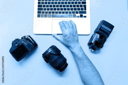 Photo camera and other accessories such as flash light and lens are on the table near laptop on background toned in blue. Man photographer prepairs photo for editing. View from the top, flat lay. photo