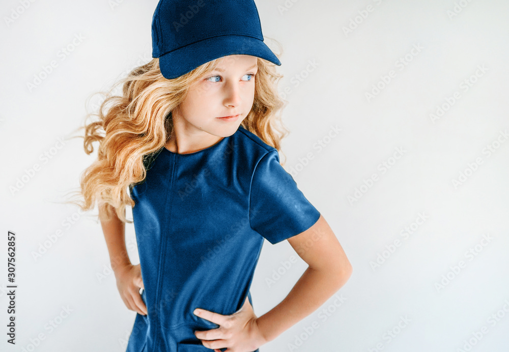 Smiling curly hair tween girl in blue leather dress and baseball cap on white background isolated Smiling curly hair tween girl in blue leather dress and baseball cap on white background isolated
