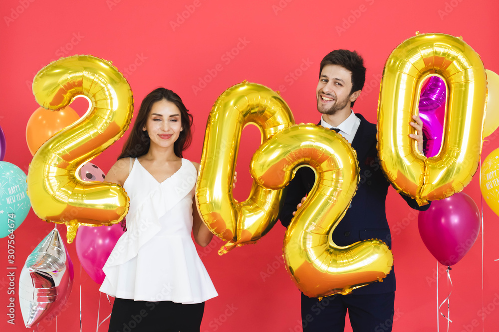 Young couple celebrating new year together, holding 2020 balloons with red backdrop.