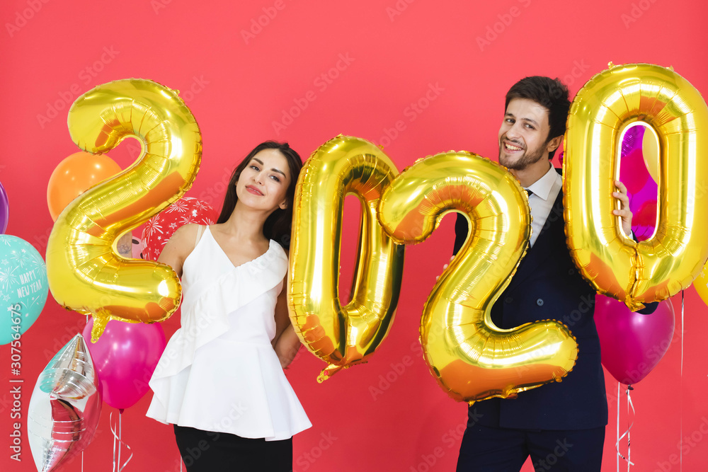 Young couple celebrating new year together, holding 2020 balloons with red backdrop.