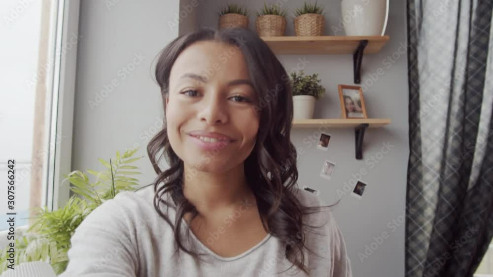 Chest-up handheld shot of vivacious young mixed race woman standing by window at home with outstretched hand and video chatting with someone, looking at camera, waving and smiling Chest-up handheld shot of vivacious young mixed race woman standing by window at home with outstretched hand and video chatting with someone, looking at camera, waving and smiling