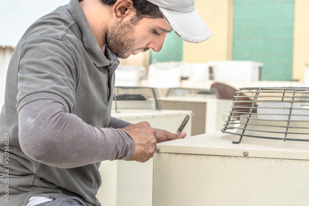 a professional electrician man is fixing the heavy air conditioner by his tools on the roof top and wearing blue color of uniform and head cap a professional electrician man is fixing the heavy air conditioner by his tools on the roof top and wearing blue color of uniform and head cap